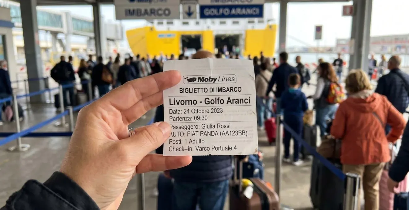 Eine Hand hält eine moderne Fährreservierungsbestätigung für die Route Livorno-Golfo Aranci, im unscharfen Hintergrund das Terminal des Hafens Livorno bei Tageslicht