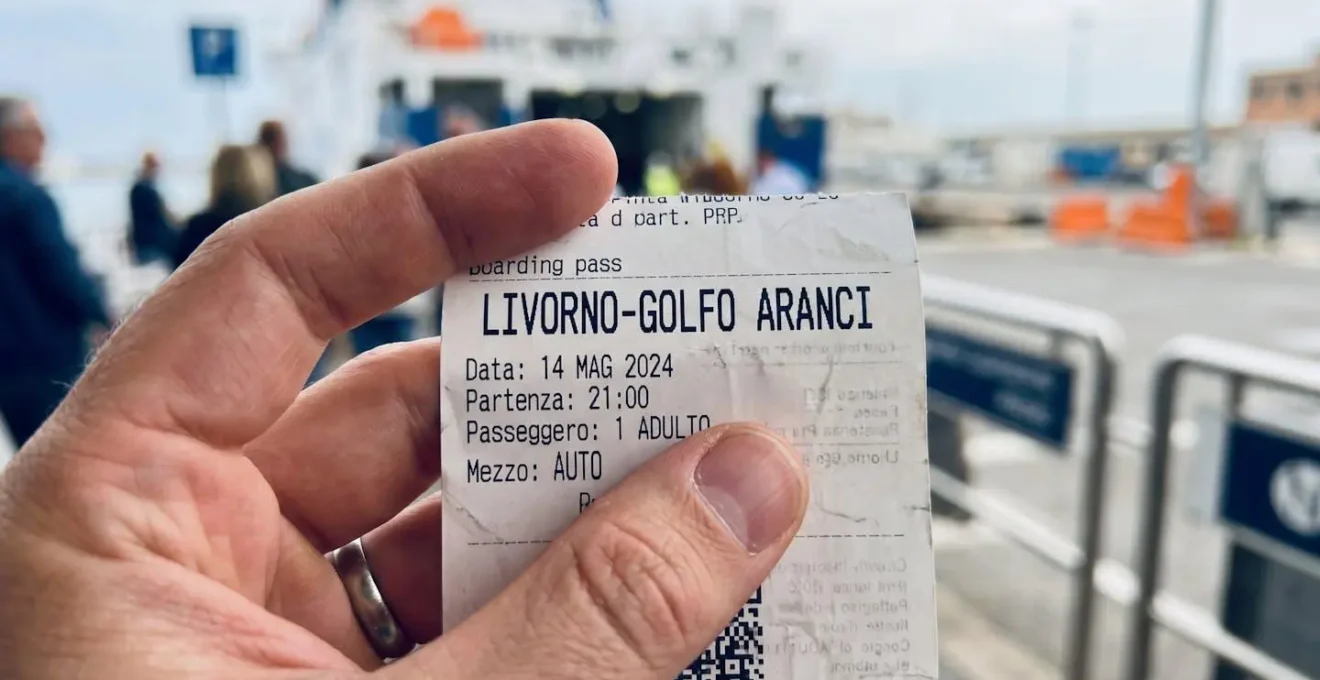 Eine Hand hält eine moderne Fährreservierungsbestätigung für die Route Livorno-Golfo Aranci, im unscharfen Hintergrund das Terminal des Hafens Livorno bei Tageslicht